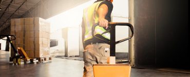 A man pulling a pallet jack toward a docking station at a warehouse. There are boxes wrapped in plastic on the equipment.