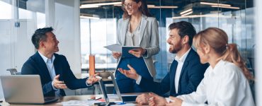 A female business professional stands in the conference room, with others sat at the table. She's presenting information.