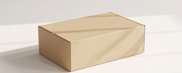 A cardboard product packaging box rests on a white table. The box has text that says "BOX MOCKUP" and "PACKAGE MOCKUP."