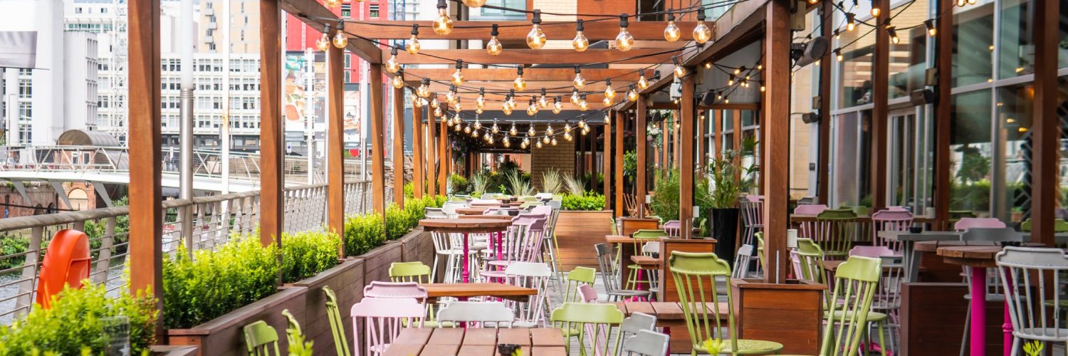 An outdoor dining patio features wooden beams with hanging string lights, wooden tables, and colorful metal chairs.