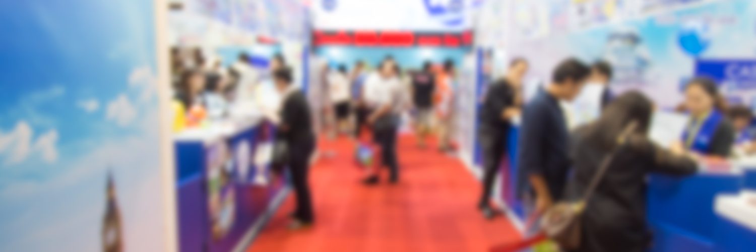 A blurred view of a crowd standing on a red carpet, surrounded by blue pop-up booths for a trade show.