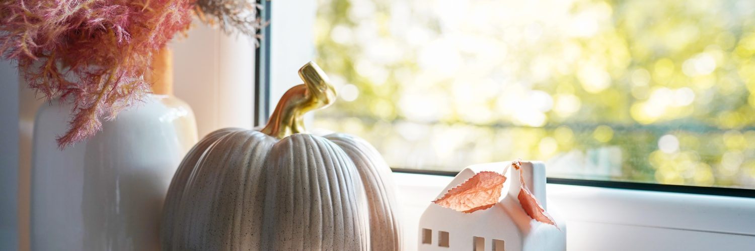 A window sill featuring an arrangement of autumn decor items, including a white vase of decorative grasses, a taupe ceramic pumpkin, and small white ceramic house.
