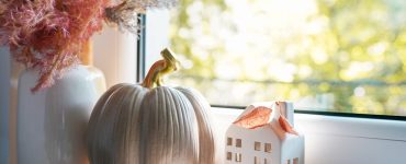 A window sill featuring an arrangement of autumn decor items, including a white vase of decorative grasses, a taupe ceramic pumpkin, and small white ceramic house.