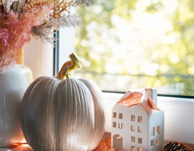 A window sill featuring an arrangement of autumn decor items, including a white vase of decorative grasses, a taupe ceramic pumpkin, and small white ceramic house.