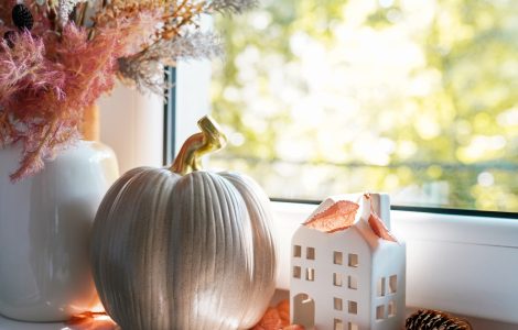 A window sill featuring an arrangement of autumn decor items, including a white vase of decorative grasses, a taupe ceramic pumpkin, and small white ceramic house.