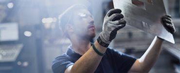 A young man wearing safety goggles and gloves holds a large sheet of metal with rectangular cutouts at a factory.