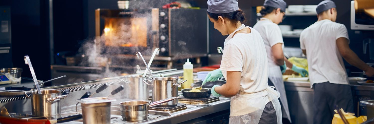 A team of cooks in the kitchen of a restaurant. They wear white shirts, aprons, and caps while working at different stations.