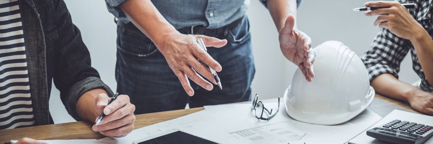 A table with construction company members standing around to discuss the blueprints. One person talks with their hands.