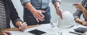 A table with construction company members standing around to discuss the blueprints. One person talks with their hands.