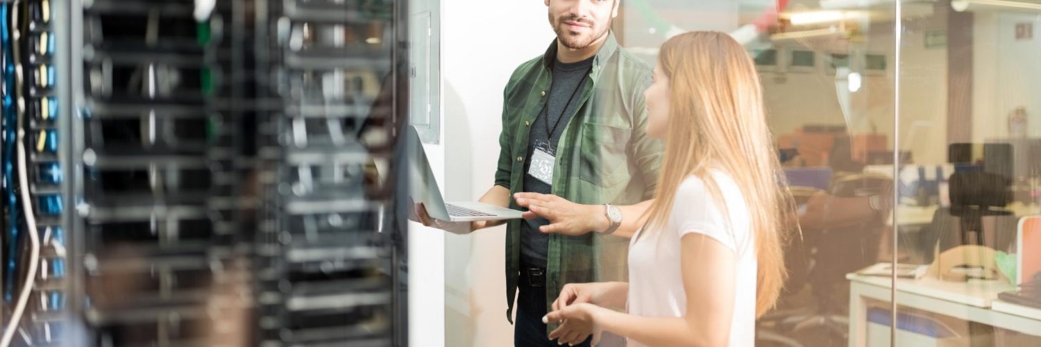 Two people stand in a server room separated from an office space by glass walls. One of them is holding a laptop.