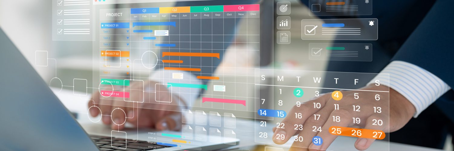 A person wearing a suit types on a laptop and touches a tablet. Two digital color-coded calendars are above the devices.