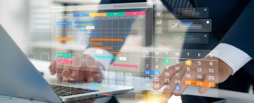 A person wearing a suit types on a laptop and touches a tablet. Two digital color-coded calendars are above the devices.
