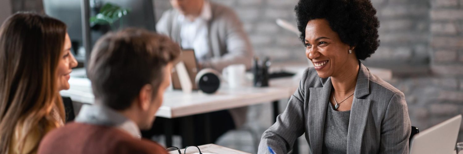 A woman in a gray blazer smiling while sitting across a desk from a man and woman. On the desk are stacks of paper and a laptop.