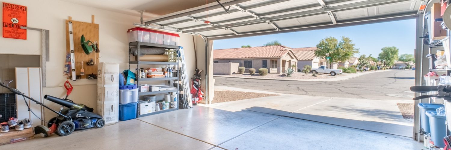 A garage with the exterior door open. There are various tools and boxes stacked against the garage wall.