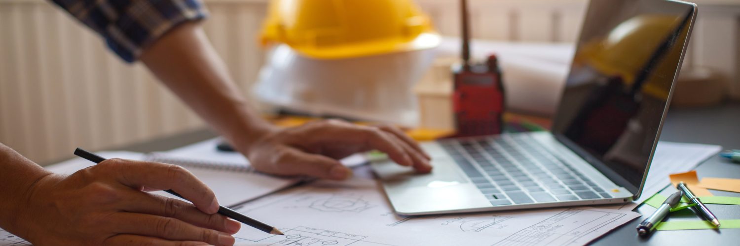 A person draws on the blueprint with one hand and uses a laptop on the other. Behind is a stack of hard hats.
