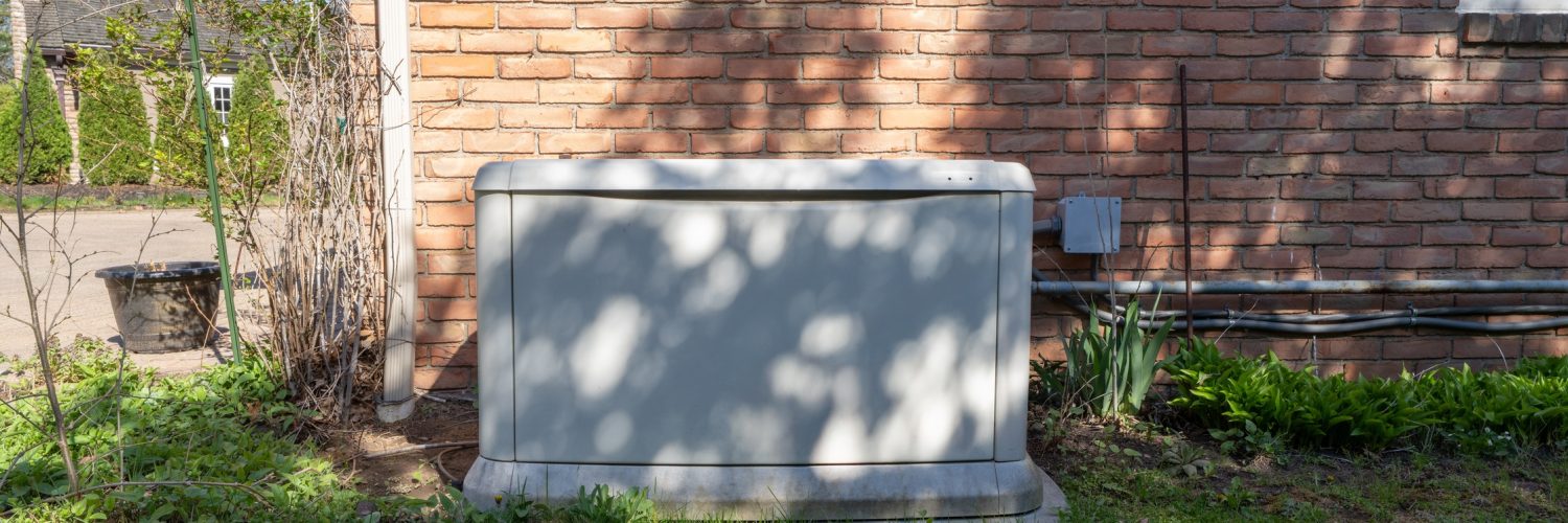 A residential backup power generator sits on a lawn in front of a brick home. A tree is casting a shadow on the generator and brick wall.