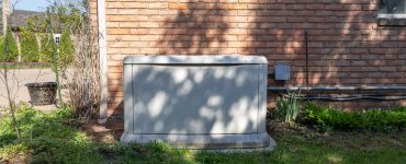 A residential backup power generator sits on a lawn in front of a brick home. A tree is casting a shadow on the generator and brick wall.