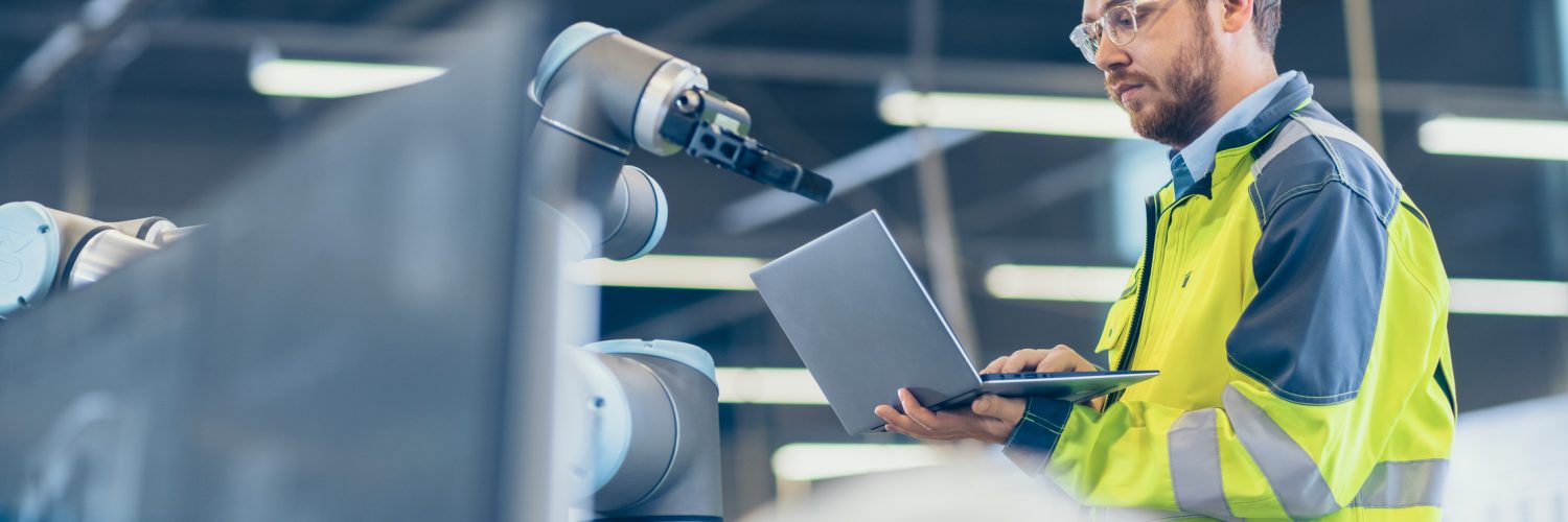 An engineer wearing a high-vis uniform uses a laptop while standing next to a robotic arm in a factory setting.