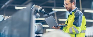 An engineer wearing a high-vis uniform uses a laptop while standing next to a robotic arm in a factory setting.