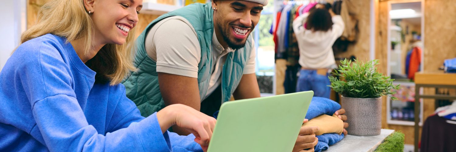 Two sales associates smile behind a green laptop in a retail pop-up shop as people shop in the background.