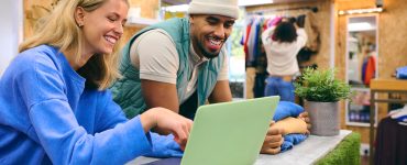 Two sales associates smile behind a green laptop in a retail pop-up shop as people shop in the background.