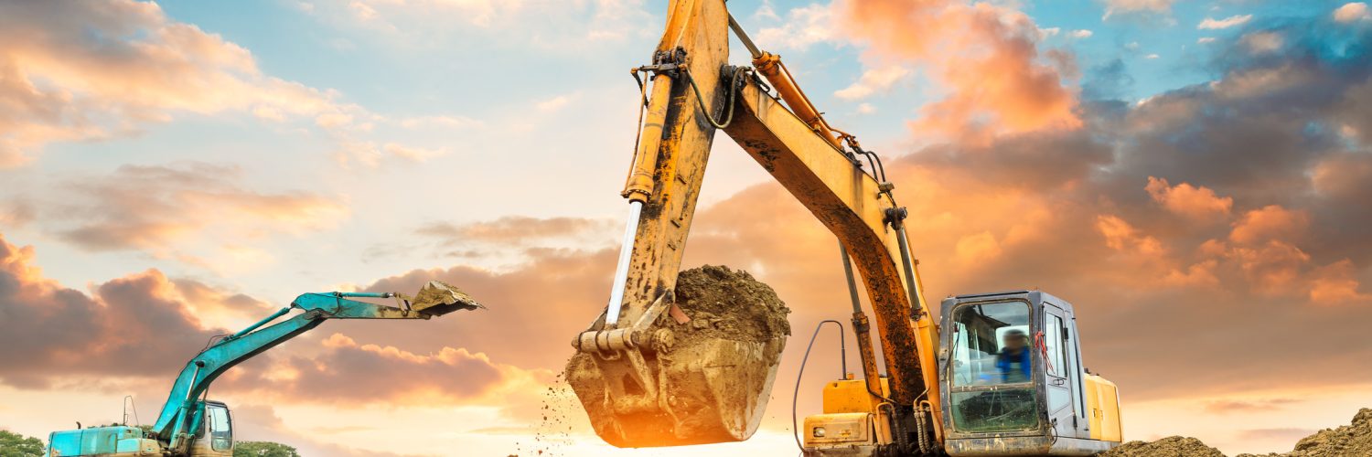 A yellow and light blue excavator working on a construction site. Behind them is the setting sun in a cloudy sky.