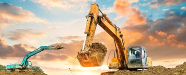 A yellow and light blue excavator working on a construction site. Behind them is the setting sun in a cloudy sky.