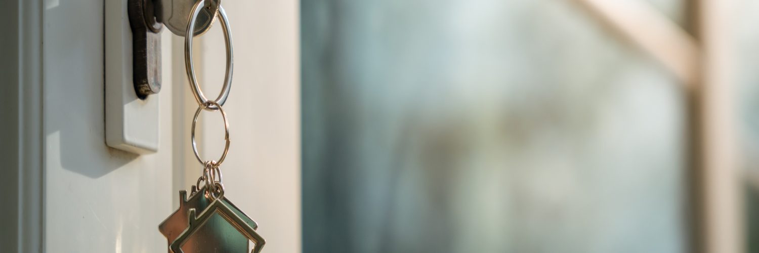 A white entrance door to a house with a key hanging from it. The key has a keychain that looks like a house.