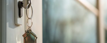 A white entrance door to a house with a key hanging from it. The key has a keychain that looks like a house.