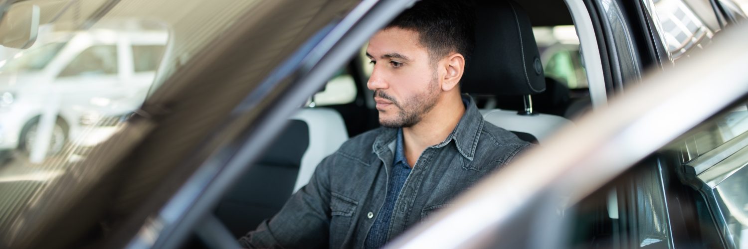 A man with a look of indifference on his face is sitting inside the driver’s seat of a car with the door open.
