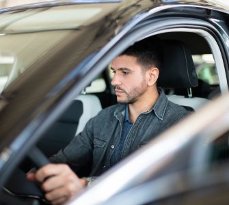 A man with a look of indifference on his face is sitting inside the driver’s seat of a car with the door open.