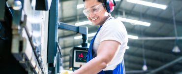 A male industrial worker wearing safety glasses and gloves is inspecting a manufactured piece in a factory.