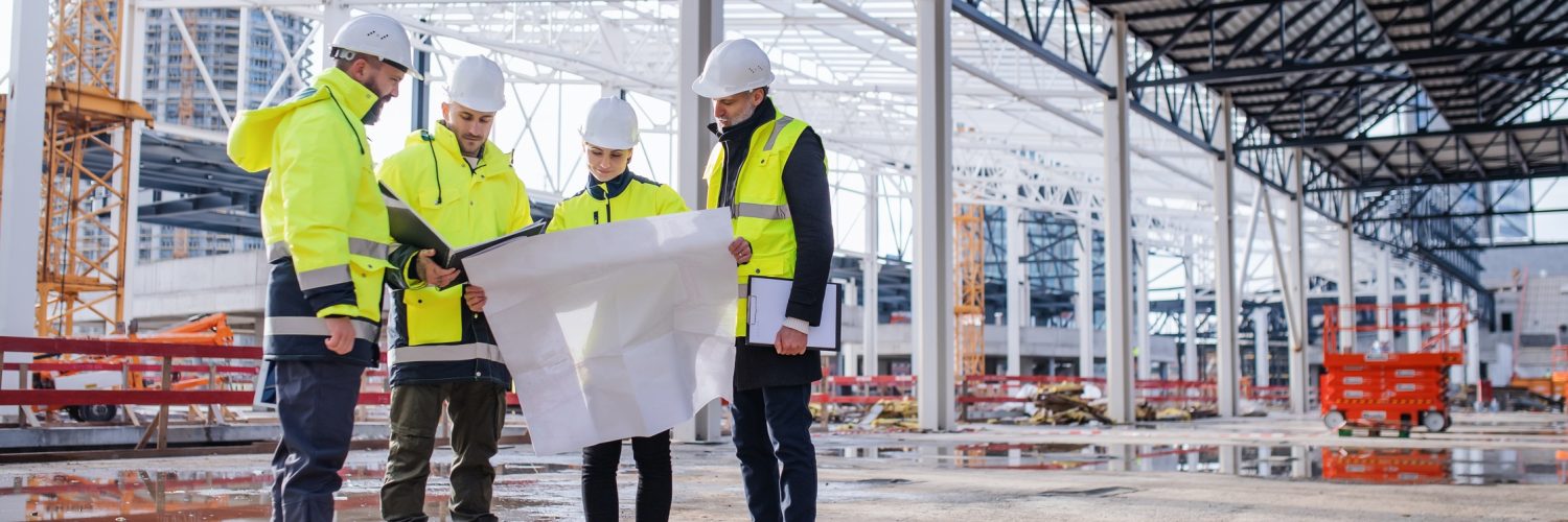 A group of engineers wearing white hard hats and safety vests huddle and look over a construction blueprint.
