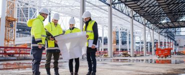 A group of engineers wearing white hard hats and safety vests huddle and look over a construction blueprint.