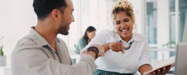 A man and a smiling woman fist-bump at a desk with a laptop and charts, while she holds an open notebook.
