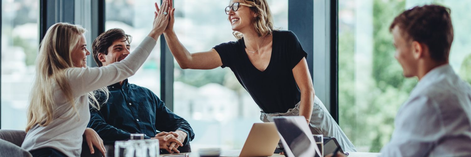 A woman standing at the head of a conference room table is high-fiving a woman sitting at the table. Two men sit at the table.