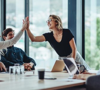 A woman standing at the head of a conference room table is high-fiving a woman sitting at the table. Two men sit at the table.