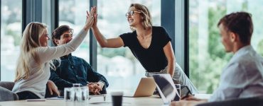 A woman standing at the head of a conference room table is high-fiving a woman sitting at the table. Two men sit at the table.