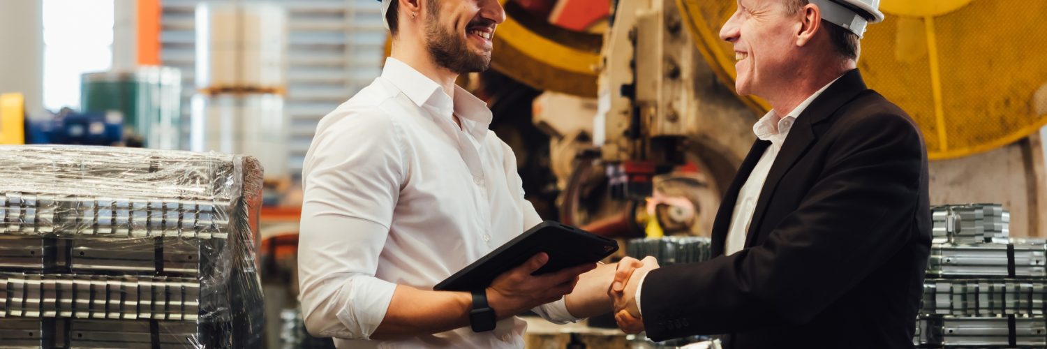 Engineer and manager wearing safety helmets shaking hands in manufacturing plant with equipment behind.