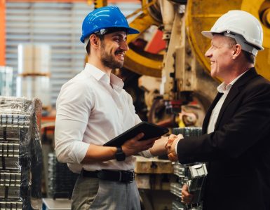 Engineer and manager wearing safety helmets shaking hands in manufacturing plant with equipment behind.