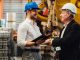 Engineer and manager wearing safety helmets shaking hands in manufacturing plant with equipment behind.
