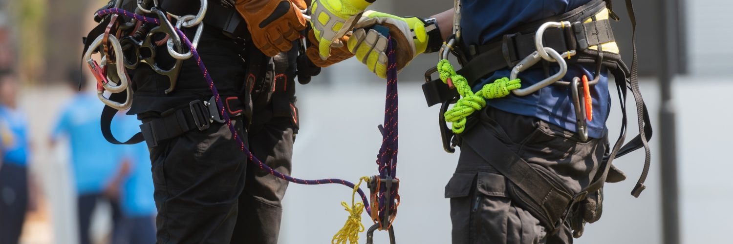 A close-up of two people wearing safety gloves and safety harnesses with ropes and multiple carabiners attached.