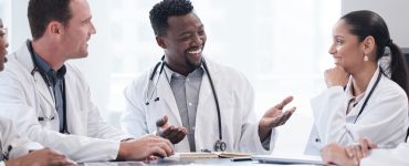 A group of doctors and healthcare staff sit around a table scattered with documents in a hospital meeting room.
